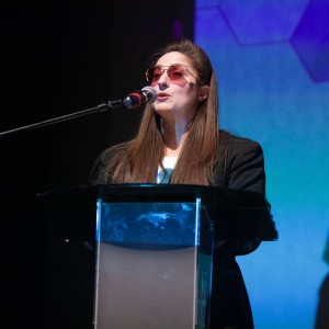 Kait Dueck is wearing a black blazer and pink-tinted sun glasses while giving a speech. Kait Is standing behind a podium and the background is black and blue.