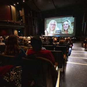 A large audience at Showplace Performance Centre watches a virtual Q&A session following the screening of Red Fever. On stage, a speaker stands at a podium, while a large screen displays Producer Ernest Webb and Co-Director Catherine Bainbridge, who are participating remotely. Webb, on the left, wears glasses and a light-coloured top, while Bainbridge, on the right, has wavy blonde hair and is smiling. Captions are visible on the screen. The dimly lit theatre highlights the stage and screen, while audience members, dressed in warm clothing, sit attentively in the darkened auditorium. The venue’s "The David Goyette Stage" sign is visible in the background.