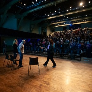 Eryn Lidster, Rodney Fuentes, and Carlotta James stand on stage as the audience gives them a standing ovation. Eryn, walking across the stage, holds a microphone out to the other two. Rodney and Carlotta stand nearby, facing her. The stage is warmly lit, contrasting with the darker seating area where the audience, dressed in various styles, is clapping. The venue features exposed beams, high ceilings, and tiered seating, creating an intimate yet spacious setting.