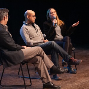 Mark Dickinson (moderator), Rodney Fuentes, and Carlotta James sit on stage discussing their film, The Monarch Ultra. Mark, on the left, wears a dark blazer and glasses, listening attentively with his hands folded. Rodney, in the centre, wears a light grey fleece and jeans, holding a microphone in his lap. Carlotta, on the right, wears a black jacket and jeans, speaking into a microphone while gesturing with her other hand. The dim stage lighting highlights the panelists against a dark background.