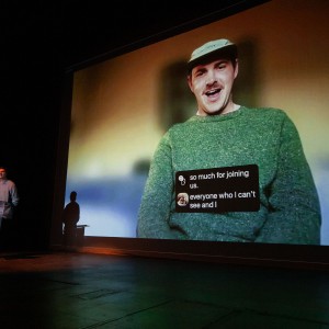 Luka Stojanovic stands at a podium on stage at the ReFrame Film Festival, moderating a Q+A while a large screen behind him displays Ben Judkins, who is participating virtually. Judkins, wearing a green sweater and a cap, is smiling while speaking about his film, The Cigarette Surfboard. Stojanovic, also wearing a sweater, is positioned to the left of the screen, illuminated by stage lighting, with his shadow cast on the wall.