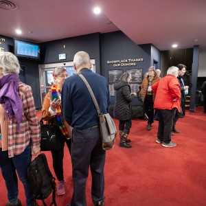 A group of people are gathered in the Showplace lobby with red carpeting. Some are engaged in conversation, while others are walking or waiting. Many are dressed in casual or semi-formal attire, with jackets, scarves, and bags. The walls feature signage, including "SUNSHINE" and "SEAN EYRE LOBBY," suggesting the name of the venue or sections within it. In the background, a doorway is open with a staff member wearing a pink shirt standing nearby. The setting appears is a theater or event space.