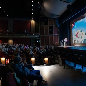 A large audience is seated in the Showplace Performance Centre, attentively listening to a speaker on stage at the ReFrame Film Festival prior to the screening of No Other Land. The stage is illuminated, with a presenter standing at a podium in front of a large screen displaying the festival's branding, including a geometric floral design and text. Captions for accessibility are visible on the screen. The auditorium, including the balcony level labeled "The David Goyette Stage," is filled with attendees, some leaning forward and others reading their programs. The dim lighting in the seating area contrasts with the brightly lit stage, drawing focus to the speaker.