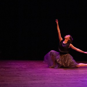 Dreda Blow performs on stage as part of the ancillary programming alongside the screening of Bound at the 2025 ReFrame Film Festival. She wears a flowing grey tulle skirt and a black sleeveless top, striking an expressive pose with one arm reaching upward and the other extended to the side. One leg is stretched behind her, toes pointed, while the other is bent beneath her. The stage is dimly lit, with warm purple and amber lighting highlighting her movement against a dark background.