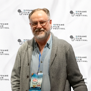 Nick van der Graaf smiles and poses in front of a white backdrop featuring the ReFrame Film Festival logo he has glasses, a beard, and short gray hair. He is wearing a light blue button-up shirt, a gray cardigan, and a blue lanyard with a VIP festival badge. The backdrop has a repeating pattern of the festival's name and a film reel icon.