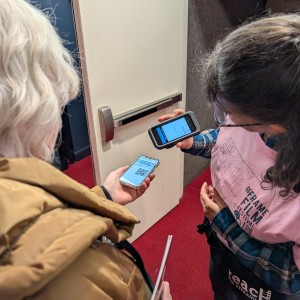 A volunteer wearing a pink ReFrame Film Festival t-shirt over a plaid shirt is scanning a QR code on another person's phone at the entrance to Showplace's auditorium. The attendee, dressed in a brown puffer jacket, holds up their phone displaying the QR code while the volunteer uses their scanner to check it. The setting features a red carpet and a white door with a push bar. The volunteer also carries a black tote bag with partially visible text reading "Reach for ReFrame."