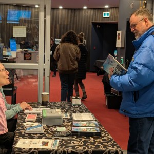 A festival volunteer wearing a pink shirt and glasses is seated behind a table covered with a black patterned cloth, engaging in conversation with another person, Nick van der Graaf, who is wearing a bright blue winter coat and is holding a program. The table is filled with programs, and small items, related to the festival. A disposable coffee cup is also on the table. In the background, a glass door reveals a hallway with other attendees walking through a red-carpeted area. The setting is the Showplace lobby.