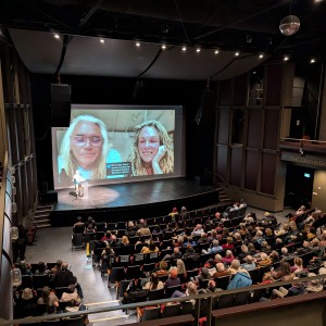 A large audience is seated in a modern theater, watching a presentation on stage. A person stands at a podium in front of a large screen displaying a video call or prerecorded video of two smiling individuals. On the left is Ernest Webb, and on the right is Catherine Bainbridge. The theater features a curved seating arrangement, dark walls, and a balcony section. Overhead lights illuminate the stage, and the audience appears attentive.