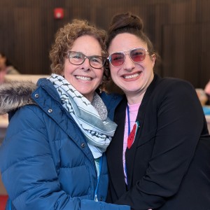 Two indviduals embrace and smile at the camera. On the left is Deborah Berrill who wears a blue winter coat and a Keffiyeh as well as glasses. On the right is Kait Dueck wearing a black blazer with a watermelon pin on right lapel. Kat has her hair in a bun and is wearing pink tinted sun glass. The atmosphere is friendly and familliar.