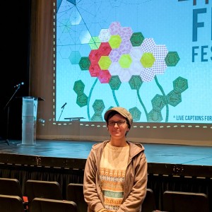 Gillian Turnham stands wearing glasses, a flat cap, a patterned sweater, and a brown zip-up hoodie in the seating area of a theater, smiling at the camera. Behind Gillian, a stage features a large screen displaying a colorful geometric quilt design with hexagonal flowers and angled lines connecting and forming an abstract shape. The words "ReFrame Film Festival" are partially visible on the screen, along with a note about live captions. A clear podium with a microphone is on the left side of the stage. The theater has black seats with red cushions.