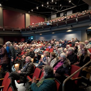 A large audience is seated in the Showplace theater with red cushioned chairs, waiting for a screening to begin. The theater has two levels of seating, with people occupying both the main floor and the balcony. The walls are deep red with dark paneling, and the ceiling features stage lighting. Some audience members are engaged in conversation, while others are looking toward the front of the theater. A few people are still finding their seats, and coats and scarves suggest a colder season. The atmosphere appears lively and anticipatory.