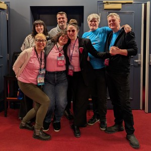 A group of seven people is posing together in a theater lobby with red carpeting and dark blue doors in the background. From left to right in the backrow is: Hannah McCammon, Michael Momtt, Michael Goede, and Philip Fox Jones. From left to right in the front row is: Melanie Buddle, Eryn Lidster, and Kait Dueck. Hannah is wearing a Keffiyeh. Michael Momtt has a grey sweater on. Michael Goede and Philip are wearing blue festival shirts. Melanie, Eryn, and Kait all have on pink festival volunteer shirts on and lanyards with Board/Staff badges. They stand close together, with some linking arms, hugging, or placing hands on each other’s shoulders. Melanie is leaning forward playfully, while Kait wearing pink-tinted sunglasses and a black blazer is reaching over holding Philip's hand. The group appears cheerful and friendly.