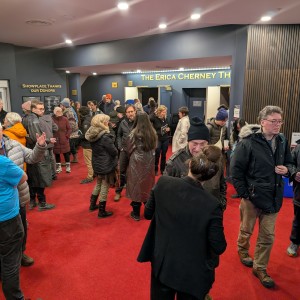 A lively crowd of people is gathered in a the Showplace lobby with red carpeting. Many attendees are dressed in warm coats, hats, and scarves, suggesting a colder season. Some are engaged in conversation, while others are walking or waiting. The illuminated sign above an entrance reads "The Erica Cherney Th," indicating "The Erica Cherney Theatre." Additional signage in the background points to "Concessions and Theatre," suggesting this is a performing arts or film venue. The space has modern decor with wood paneling and recessed lighting.