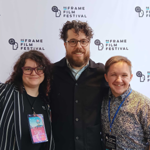 Three people are posing together in front of a white backdrop featuring the "ReFrame Film Festival" logo, which includes a stylized film reel and text. They are smiling and standing closely. The person on the left, ReFrame Creative Director Eryn Lidster has curly dark hair, wears purple glasses, a black-and-white striped blazer, and a staff badge labeled "Creative Director." The person in the middle, Filmmaker Rob Viscardis, has curly hair, a beard, and wears glasses, a black jacket, and a light blue shirt. The person on the right, Filmmaker Kurtis Watson, has short hair, wears a floral-patterned button-up shirt, and has a festival badge on a blue lanyard. The lighting is bright and even, clearly illuminating their faces.