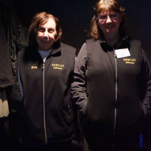 Showplace Volunteers Marg and Patti stand side by side in a dimly lit area, both wearing matching black vests with "SHOWPLACE Volunteer" embroidered in yellow. Marg, on the left, has short brown hair and a warm smile, while Patti, on the right, wears glasses and has shoulder-length brown hair with a name tag pinned to her vest. The dark background and lighting suggest they are assisting with the ReFrame Film Festival during a screening.