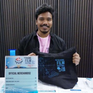 Volunteer Praveen Prakash Koottuparambil is standing behind a merchandise table at ReFrame Film Festival, smiling and holding up a black tote bag with the festival's 2025 branding in blue. He is wearing a pink festival t-shirt under a black shirt. The table in front of him displays various official ReFrame merchandise items, including folded t-shirts and other tote bags. A sign on the table lists merchandise prices. The background features a dark wooden slat wall.