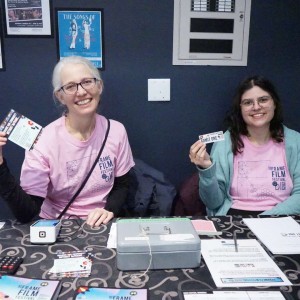 Two smiling volunteers, Jennifer VanLeeuwen (left) and Kate Long (right), sit behind the ticketing table at ReFrame Film Festival's Showplace location, both wearing matching pink festival t-shirts. Jennifer, who has glasses and white hair, holds up a promotional flyer, while Kate, also wearing glasses and a light blue cardigan, holds an "ADMIT ONE" ticket. The table is covered with a black and white patterned cloth and contains a cash box, flyers, a calculator, a card reader, and other festival materials. The background features a dark blue wall adorned with posters.