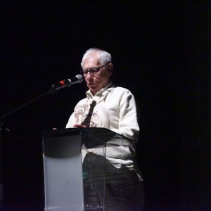 Jim Able stands at a clear podium on a dimly lit stage, speaking into a microphone. He has glasses and white hair and is wearing a light-colored button-up shirt Jim appears focused as he addresses the audience. There is dark background and a spotlight on him as he address an audience at the **ReFrame Film Festival**.