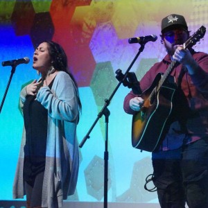 Missy Knott and Nathan Miller perform on stage, illuminated by colorful lighting from a large screen displaying abstract hexagonal patterns. Missy is on the left wearing a black dress and a light gray cardigan. She sings passionately with one hand on her chest. Nathan is on the right wearing a baseball cap and a maroon shirt while playing an acoustic guitar and singing into a microphone. The stage setting and vibrant background suggest a live musical performance at the **ReFrame Film Festival**