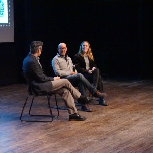 Three people are seated on a stage, engaged in a panel discussion. The person on the left facing the other two, is moderator Mark Dickenson who is dressed in a dark suit. Rodney Fuentes is in the middle wearing glasses and a light gray sweater. Rodney is speaking, while to the right, Carlotta James dressed in black, listens attentively with a slight smile. Part of a large screen with a QR code is visible in the background. The wooden stage floor and dark surroundings create an intimate setting as they address an audience at the **ReFrame Film Festival**.