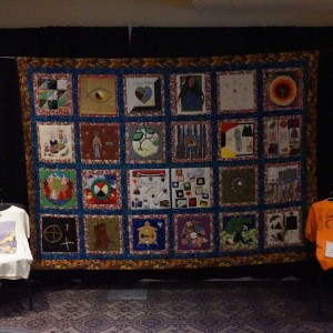 A large, handmade quilt is displayed against a black backdrop, featuring 20 individual squares, each with unique designs, symbols, and imagery. This is A group quilt project by **Kawartha Truth and Reconciliation Support Group** members on the legacy of residential schools. The quilt is bordered by a colorful patterned fabric. On either side of the quilt, chairs hold t-shirts with printed designs, one white and one orange with the words "Every Child Matters" on it. A red cloth is draped over a small table to the right, accompanied by a stack of papers or booklets.