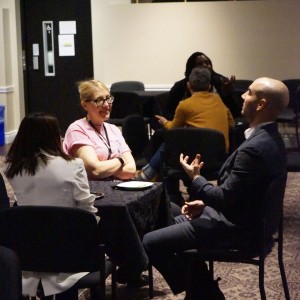 A small group of people is engaged in conversation at a table covered with a black cloth in a dimly lit room. Jose Piranian is in a suit gesturing animatedly while speaking, while Melanie Buddle, wearing glasses and a pink shirt, listens with her arms crossed. In a white blazer, Simal Iftikhar sits with their back to the camera. In the background, other groups are having discussions, and a sign on the wall reads **"COGECO | STUDIO,"**.