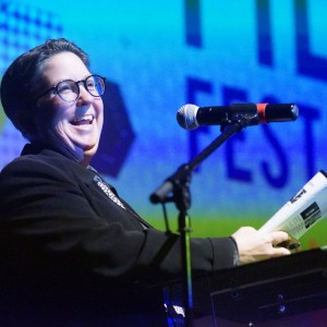 Karleen Pendleton is wearing glasses and a black suit while standing at a clear podium, speaking into a microphone with a broad smile. Karleen is holding a rolled-up document or program in one hand. The background features a large screen with colorful hexagonal patterns and partially visible text, including the words **"FILM FESTIVAL."** The stage lighting casts a vibrant glow, emphasizing the speaker’s enthusiastic expression, they are presenting an event at a film festival.