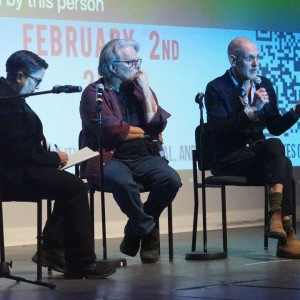 Three people are seated on a stage, engaged in a panel discussion. The moderator Karleen Pendleton Jimenez is on the left dressed in dark clothing, and holding a notepadThe middle person, Mike Munn, wearing glasses and a maroon jacket, is listening attentively with their arms crossed. Michael Mabbott is on the right, dressed in a dark suit with tan boots, and is speaking into a microphone while gesturing with their hand. Behind them, a large screen displays text, including "FEBRUARY 2ND" and a QR code. A disposable coffee cup is on the floor near the speaker. They are addressing an audience at **ReFrame Film Festival**.