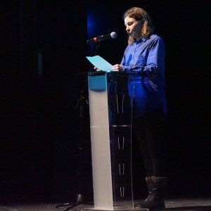 A person stands at a clear podium on a dimly lit stage, reading from a sheet of paper while speaking into a microphone. They are wearing a loose-fitting blue shirt, dark pants, and boots. A spotlight highlights them against the dark background, emphasizing their presence. They are addressing an audience at **ReFrame Film Festival**.