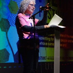 Jay Simmon stands at a clear podium, speaking into a microphone while holding a few sheets of paper. They have curly gray hair, glasses, and are wearing a pink t-shirt over a long-sleeved black shirt with dark pants. The background features a large screen displaying colorful geometric designs and partially visible text. The lighting highlights the speaker against the darker stage, as they are addressing an audience at **ReFrame Film Festival**.