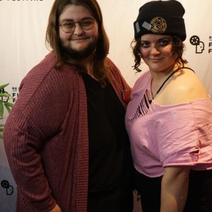 Two individuals pose in front of a white background with the words "ReFRAME FILM FESTIVAL" printed several times across. On the left is board member Angeni Lovelady wearing glasses, a burgundy cardigan with a black shirt underneath, and a watermelon pin. Beside Angeni on the right is Eryn Lidster. Eryn is wearing a black beanie with an older yellow ReFrame logo, a pink shirt that has been cut up and a black skirt. Eryn also has black eye makeup and white gloves on. This is from the costume contest.