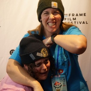 Two people pose playfully in front of a "ReFrame Film Festival" backdrop. Lindsey Unterlander, in the blue shirt, wearing a black beanie, has a wide, exaggerated grin while putting the other person, Eryn Lidster, in a lighthearted headlock. Eryn, who is in a pink shirt and a dark blue beanie, leans forward, making a comical expression. Both are wearing festival lanyards, suggesting they are attendees or staff. Their interaction appears friendly and fun, capturing a candid, humorous moment at the event.