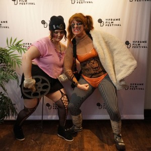 Two people in creative, costume-style outfits pose energetically in front of a ReFrame Film Festival backdrop. Eyrn Lidster, on the left, wears a pink cut-up top, black shorts, fishnet stockings, a black beanie, and goggles resting on their forehead while holding a large film reel prop. Naomi DuVall, on the right, wears an orange and black bodysuit with a corset-like design, a white fur-lined coat draped over one shoulder, and bold eye makeup. Both have bold face paint or makeup and are crouching slightly in dynamic poses. A potted plant is visible to the side, and the wooden floor adds warmth to the setting. Their playful expressions suggest a fun, themed event or cosplay element at the festival.