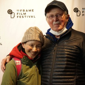 Two people pose together in front of a white backdrop featuring the ReFrame Film Festival logo. The person on the left is wearing a green jacket, a beige knit hat, and a red backpack, smiling warmly. The person on the right, who has an arm around them, is wearing a black puffer jacket, a navy blue cap, glasses, and a partially lowered face mask. They both appear happy, dressed for cold weather, and are likely attending the festival.