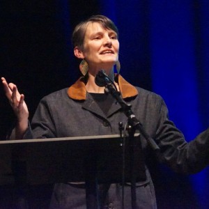 Kate Alton stands at a podium, speaking into a microphone with their arms raised expressively. They are wearing a dark coat with a brown collar and large leaf shaped earrings. The background is dark with blue stage lighting, suggesting a formal or theatrical event. Their facial expression conveys passion and engagement.