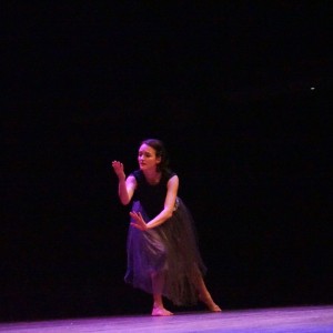 Barefoot, Dreda Blow, performs on stage in a black top and a flowing semi-transparent grey skirt. She is in a dramatic pose, with one hand reaching forward and the other extended downward, as if expressing deep emotion through movement. Her facial expression conveys intensity and focus. The stage lighting casts a soft glow on her, while the background remains dark, emphasizing her presence.