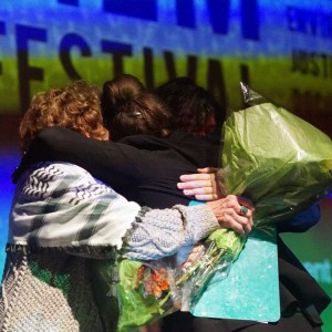 Deborah Berrill, Kait Dueck, and Eryn Lidster (left to right) embrace on stage. Deborah, wearing a Keffiyeh, holds a bouquet of flowers in one hand. Behind them a large screen displays the partially visible word "FESTIVAL" with other words such as "ENVIRONMENTAL" and "JUSTICE" to the right. The be background is colorful and abstract.