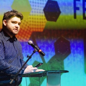 Lucas Graham is standing at a clear podium, speaking into a microphone while holding a phone in one hand. He has short, wavy brown hair and a beard and is wearing a dark blue, patterned button-up shirt. Behind him, a large screen displays colorful abstract shapes, hexagonal patterns, and partially visible text, including part of the word "FESTIVAl" and fragments of a sentence. The lighting highlights his face. He is on stage at a screening at Showplace.