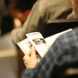 The shoulder of a person wearing a blue plaid shirt as they are seated in an auditorium, holding and reading a program. The text and images on the program are partially visible but not fully legible. The background is blurred, showing other attendees seated in rows of chairs.