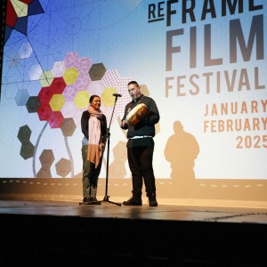 Destiny Cardinal and James Mixemong perform on stage at the Opening Night event of the ReFrame Film Festival on January 23, 2025. Destiny stands at a microphone, wearing a scarf and denim pants, smiling warmly. James, dressed in black, holds a hand drum and appears to be singing and playing. The large screen behind them displays the ReFrame Film Festival branding, including geometric floral artwork and event dates. Their shadows are cast on the screen, adding depth to the scene. The lighting highlights the performers against the dark stage surroundings.