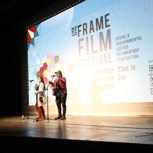 Missy Knott (Singing Wild Rice Girl) and Nathan Miller perform on stage during the Opening Night event of the ReFrame Film Festival on January 23, 2025, kicking off the festival. Missy, standing at a microphone, sings while Nathan, wearing a beanie and a red jacket, plays an acoustic guitar. The large screen behind them displays the ReFrame Film Festival branding, including geometric floral artwork, event dates, and a QR code. Their shadows are cast on the screen, adding depth to the scene. A water bottle sits on the stage near Missy's feet. The lighting highlights the performers against the dark stage surroundings.