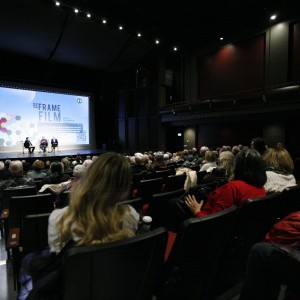 A large audience is seated in the Showplace Performance Centre, attentively watching a panel discussion on stage at ReFrame Film Festival. Three speakers sit on stage in front of a large screen displaying the festival’s branding, including a geometric floral design and text. The auditorium is dimly lit, with the stage as the primary light source. The audience, dressed in warm clothing, is engaged, with some leaning forward, holding coffee cups, or taking notes. The venue’s balcony, labeled "The David Goyette Stage," is visible in the background, with a few attendees seated above.