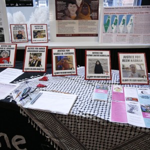 A table partially covered with a black-and-white keffiyeh is set up in the lobby before a screening. Various pamphlets, flyers, and framed photographs with red borders are displayed, featuring images of Palestinian children who have been murdered. Alongside there is text related to justice and human rights. Some materials specifically mention "Justice for" the names of the children with the text "Glory to Our Martyrs" underneath. There are also sign-up sheets with pens and watermelon fabric pins on the table. In the background, posters with additional information and images about Palestine are mounted on a window or wall. A can of soda is visible on the right side of the table.