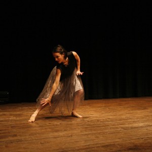 Dreda Blow performs on stage as part of the ancillary programming alongside the screening of Bound at the 2025 ReFrame Film Festival. She wears a flowing grey tulle skirt and a black sleeveless top, striking a dynamic pose with one arm extended downward and the other bent behind her. Her knees are bent, and she leans forward with an intense, focused expression. The dimly lit stage and dark curtain backdrop emphasize her movement, while soft lighting highlights her form against the wooden floor.