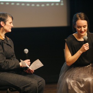 Kate Alton, incoming Executive Director of Public Energy Performing Arts, and Dreda Blow, performer and retired principal ballerina, sit on stage engaged in conversation and Q+A. Kate, on the left, wears a dark denim-like outfit with a brown collar, holding a microphone and a sheet of paper while attentively listening. Dreda, on the right, wears a black sleeveless top and a voluminous grey tulle skirt, speaking into a microphone while looking down. The dimly lit background features a large projection screen.