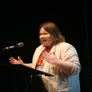 Heather Ray stands at a podium, speaking into a microphone with an expressive gesture. She wears a light-colored blazer over an orange top with a lanyard and festival badge. The dimly lit background contrasts with the stage lighting, which highlights her face and upper body. Her engaged expression suggests she is delivering a speech or presentation.