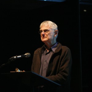 Alan Buchkowski stands at a podium, speaking into a microphone. He wears a dark zip-up jacket over a collared shirt and glasses. The lighting highlights his face against a dark background, creating a focused and intimate atmosphere. A small object rests on the podium near his hand. The setting suggests a formal speech or presentation.