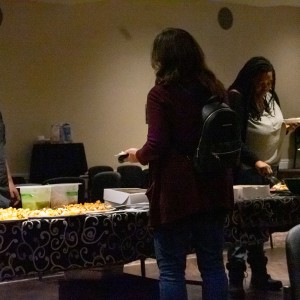 Four people stand around a buffet table covered with a black and white patterned tablecloth. The table is filled with various appetizers and snacks on colorful plates, along with beverage cans and napkins. On the right facing the camera, Martin from La Mesita Restaurant is wearing a dark blue shirt as he speaks to a woman in a dark red sweater wearing a black back. Her back is to the camera but she appears to be holding a plate of food. The other two attendees stand on either side of the table serving themselves food. The background features a small round table covered by a black tablecloth, and a fifth person who is only partially visible.