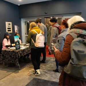 Two volunteers in pink shirts sit behind the ticketing table at ReFrame Film Festival's Showplace location. One volunteer is wearing a black baseball hat while the other is obscured by a festival sign. The table is covered with a black and white patterned cloth and contains festival materials. A line of five people wait to buy tickets, some are dressed in warm coats, hats, and scarves, suggesting a colder season. The picture is taken from the back of the line with the volunteers in the back left and the line extending to the front right. The background features a dark blue wall adorned with posters.