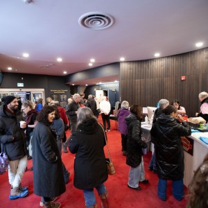 An engaged crowd of people is gathered in a the Showplace lobby with red carpeting. Many attendees are dressed in warm coats, hats, and scarves, suggesting a colder season. Some are engaged in conversation, while others are interacting with volunteers at the Merch and table. The space has modern decor with wood paneling and recessed lighting.